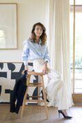 Une femme est assise à moitié sur un tabouret en bois. Elle porte des talons noirs, un pantalon ample blanc à fines rayures noires, et une chemise bleu ciel. Dans sa main droite, elle tient un blazer bleu marine, posé le long du tabouret. Derrière elle, une baie vitrée et un tableau abstrait en noir et blanc complètent le décor.