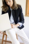 Une femme écrit de la main gauche sur un bloc-notes aux feuilles blanches. Elle porte un pantalon blanc à fines rayures noires, une chemise bleu ciel et un blazer bleu marine. Assise sur un tabouret en bois, elle regarde ce qu’elle est en train d’écrire. La photo est prise de face.