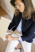 Une femme écrit de la main gauche sur un bloc-notes aux feuilles blanches. Elle porte un pantalon blanc à fines rayures noires, une chemise bleu ciel et un blazer bleu marine. Assise sur un tabouret en bois, elle regarde ce qu’elle est en train d’écrire. La photo est prise en plongée.