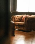 Un fauteuil en velours marron capitonné est placé dans une pièce au parquet en bois, baignée par une lumière douce provenant d'une grande fenêtre voilée. Un coussin rouge repose sur le fauteuil, ajoutant une touche de chaleur. Une table basse ancienne en bois est partiellement visible au premier plan, floutée, suggérant une atmosphère intime et feutrée. L’image dégage une sensation de calme, de confort et de nostalgie, évoquant un intérieur classique et élégant.