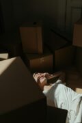 Une personne allongée sur le sol, entourée de nombreuses boîtes en carton, probablement en plein déménagement. Elle porte un t-shirt blanc et semble fatiguée ou stressée, les mains couvrant partiellement son visage. Une lumière naturelle provenant d'une fenêtre éclaire partiellement son corps et les boîtes autour d’elle, créant un contraste marqué entre l’ombre et la lumière. L’atmosphère suggère une sensation d'épuisement ou de surcharge.